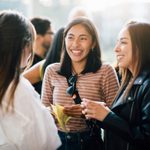 Three young women smile and chat together at an indoor social gathering or event.