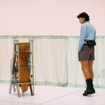 An adult walking around and looking at a stand-alone art piece, surrounded by sheer green low-hung curtain