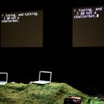 Two laptops are placed on a grassy surface, facing each other. Behind the laptops, two screens that display the text, 