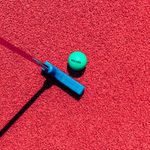 A green golf ball with the word WALKER printed in black, atop red turf grass next to a blue golf putter.