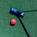 A red golf ball with the word WALKER printed in black, atop a turf grass next to a blue golf putter.