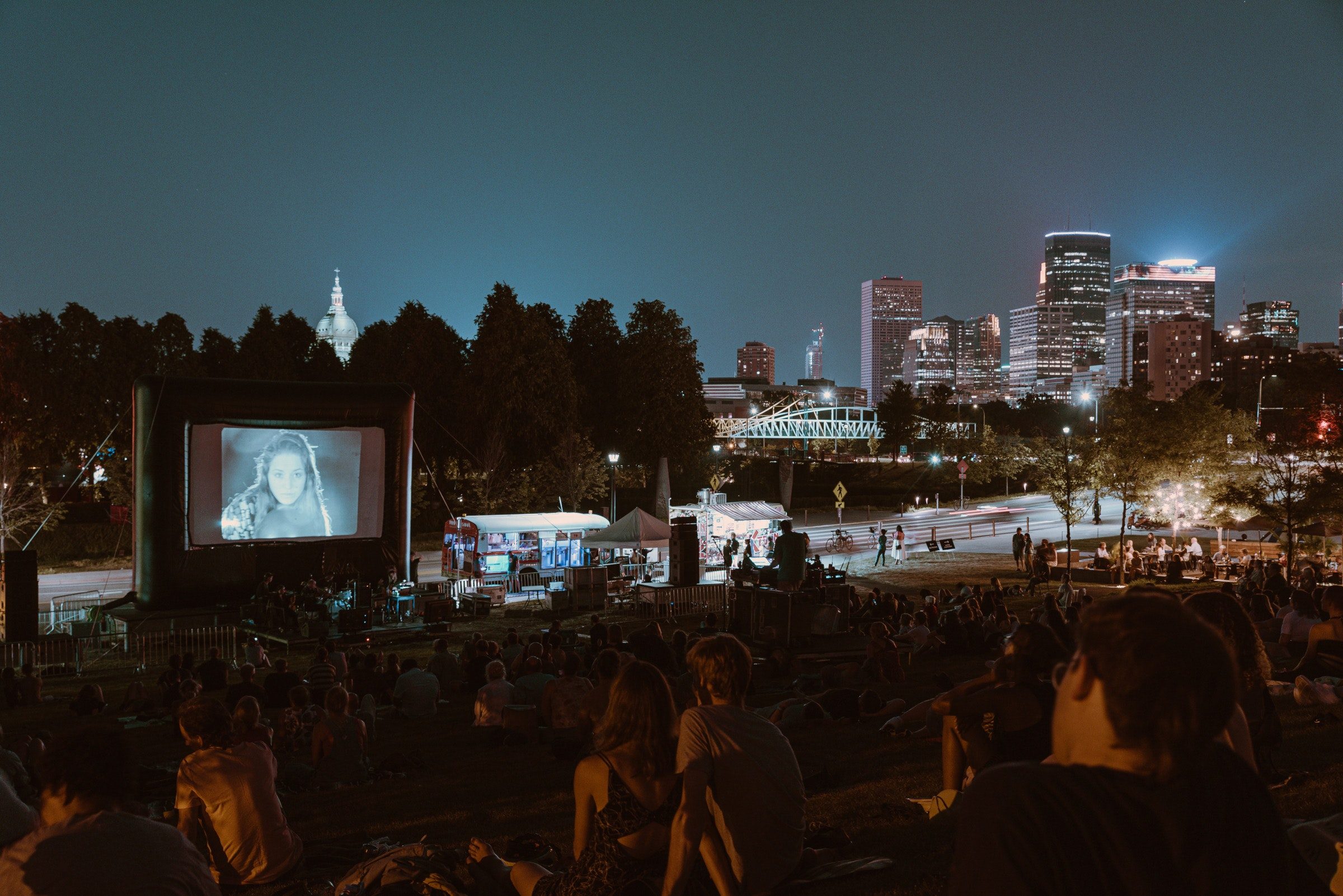 Crowds of people on pinic blankets on a hillside next to a museum watch a film projected on a large outdoor screen with a band playing in front of it and the Minneapolis skyline in the background.