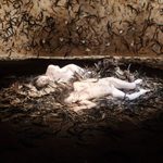 Live art installation of two adults painted in white and grime, laying on a pile of natural debris
