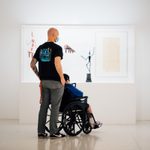 One standing masked adult with a seated adult in a wheelchair look at a photo on the wall, surrounded by other art pieces.