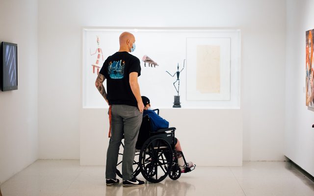 One standing masked adult with a seated adult in a wheelchair look at a photo on the wall, surrounded by other art pieces.