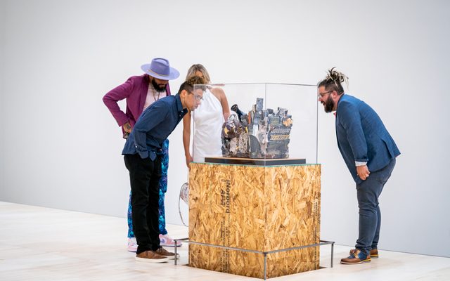 4 adults look closely at a recycled-materials art installation in a glass case, atop a particle wood stand.