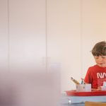 A young adolescent wearing a red NASA tshirt, holding a paintbrush and looking at art supplies on a table