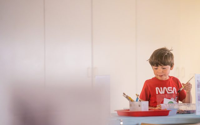 A young adolescent wearing a red NASA tshirt, holding a paintbrush and looking at art supplies on a table
