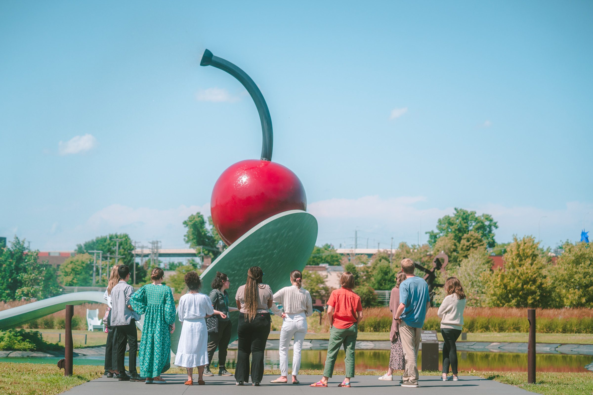 Very large outdoor sculpture of a cherry on a spoon next to a body of water at a park with grass, with a dozen people looking and admiring it.