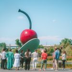 Very large outdoor sculpture of a cherry on a spoon next to a body of water at a park with grass, with a dozen people looking and admiring it.