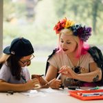 peli2024ffs0601_032 An adult with blonde and pink hair and a flower crown is sitting at a table working on art with a child wearing a backwards baseball cap