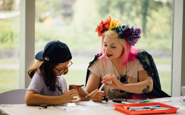An adult with blonde and pink hair and a flower crown is sitting at a table working on art with a child wearing a backwards baseball cap