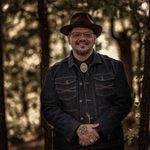 An adult with medium brown skin stands looking at the viewer in a forest. They are wearing a brown fedora and jean jacket.