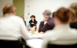 A group of adults sit and listen in a conference room at a table filled with papers and beverages
