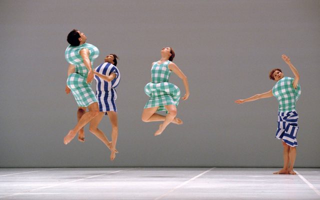 3 dancers jumping in place and one dancer posed on stage wearing green plaid and blue striped garments