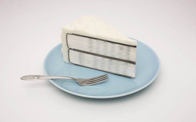 An art piece of a sliced white cake filled with two cut books that show the binding and pages, atop of a blue porcelain plate next to a fork