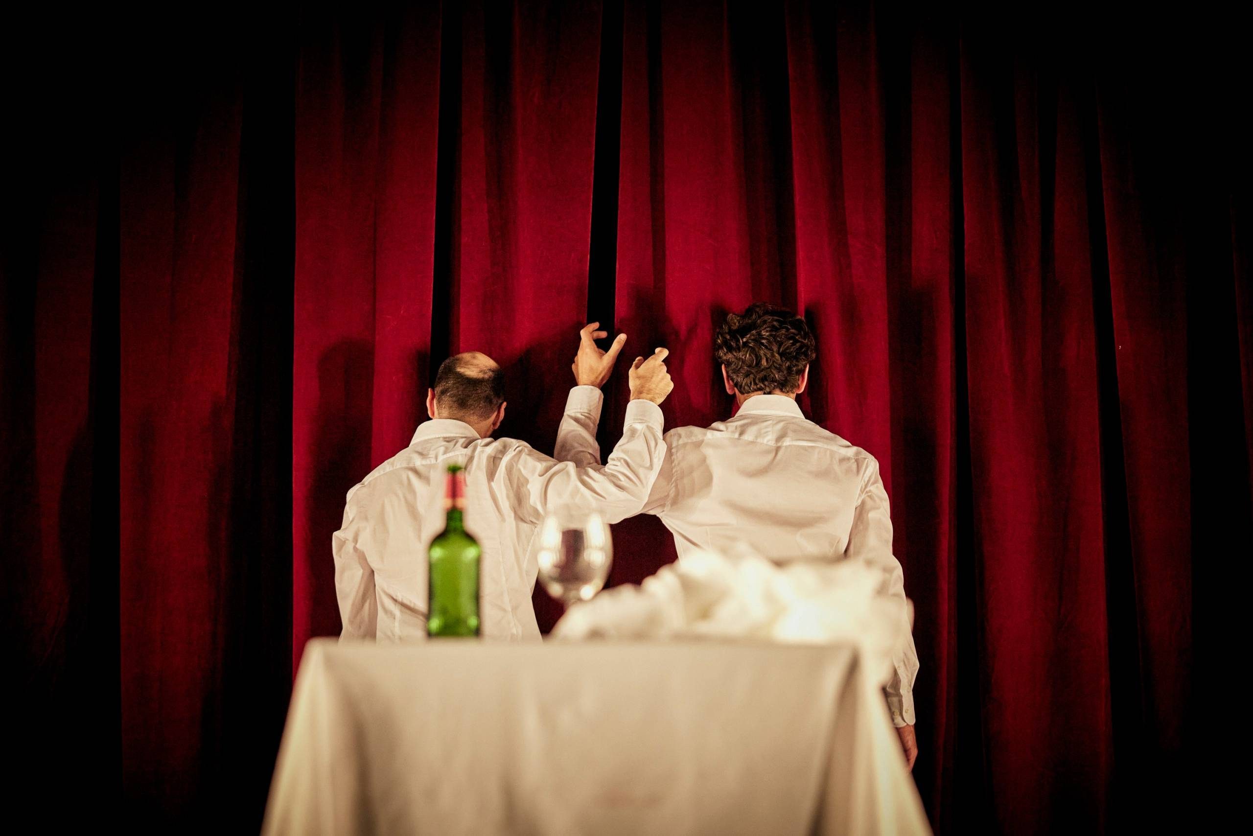 Two light-skinned performers stand with their backs to a table with a wine bottle, glass, and tablecloth.
