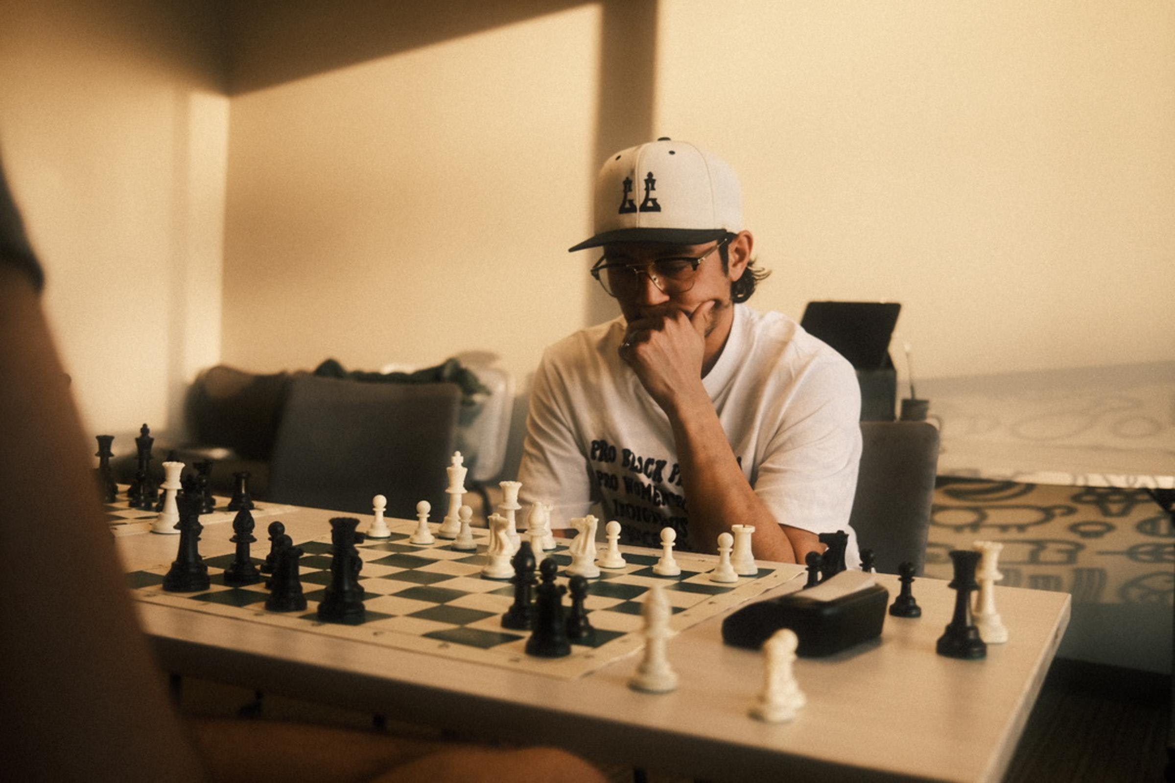 A person wearing glasses and a hat with a chess piece design is intently focusing on a chessboard during a game in a softly lit room. There are black and white game pieces all over the table.