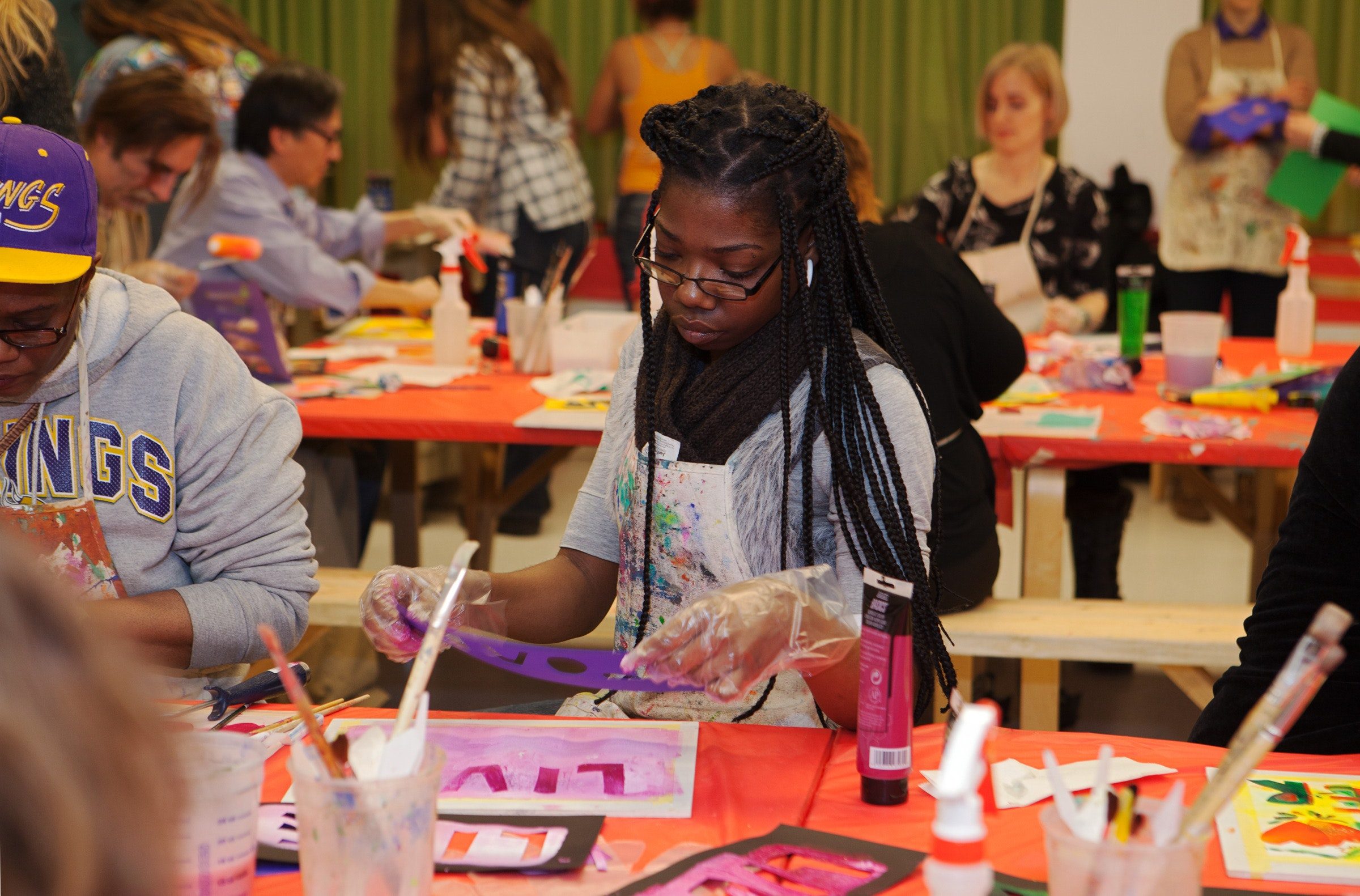 A woman paints at a crowded table during an art class, surrounded by art supplies and other participants.