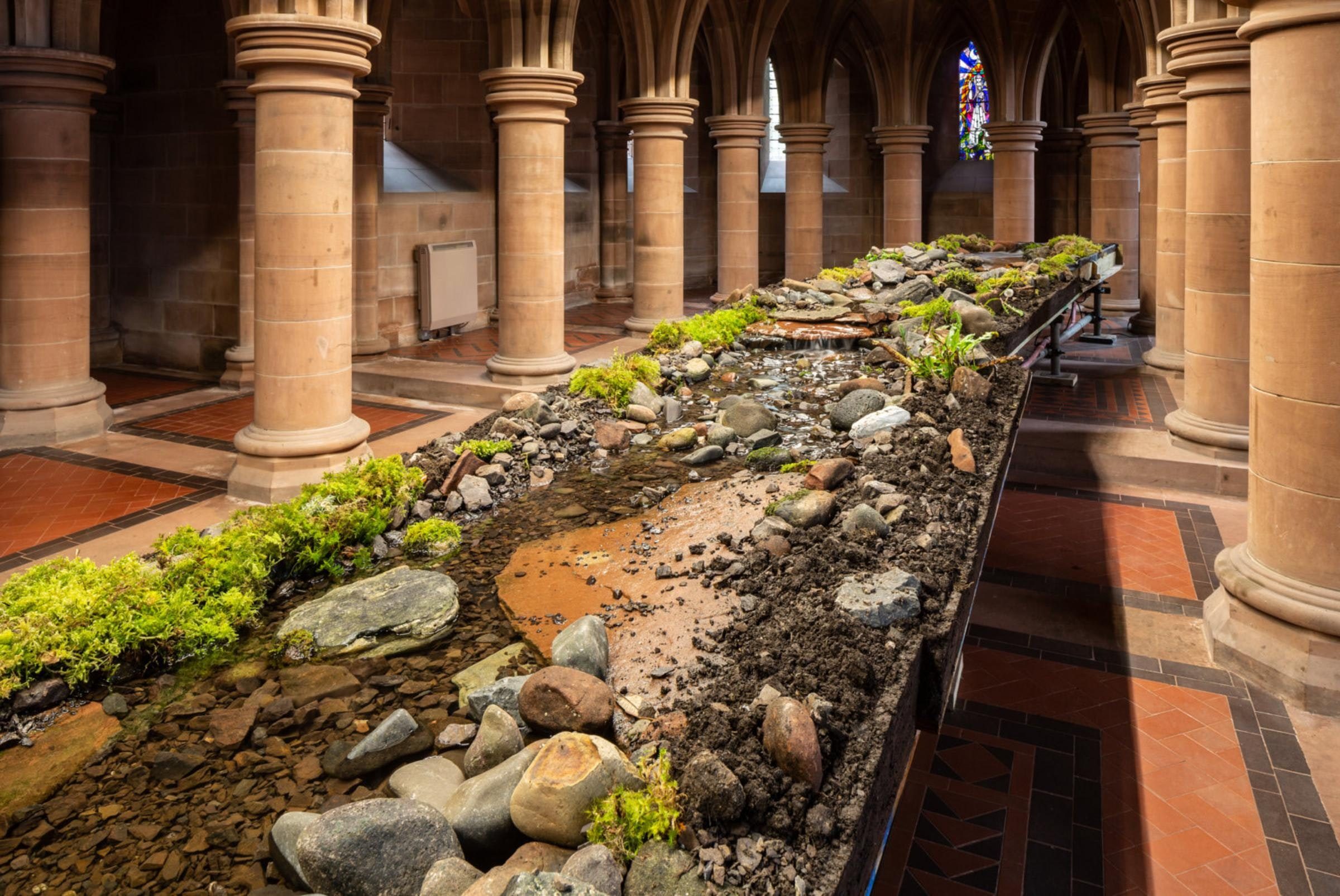 A large room with redbrown circular columns surrounding a long table, covered in soil, rocks, green plants, and running water.