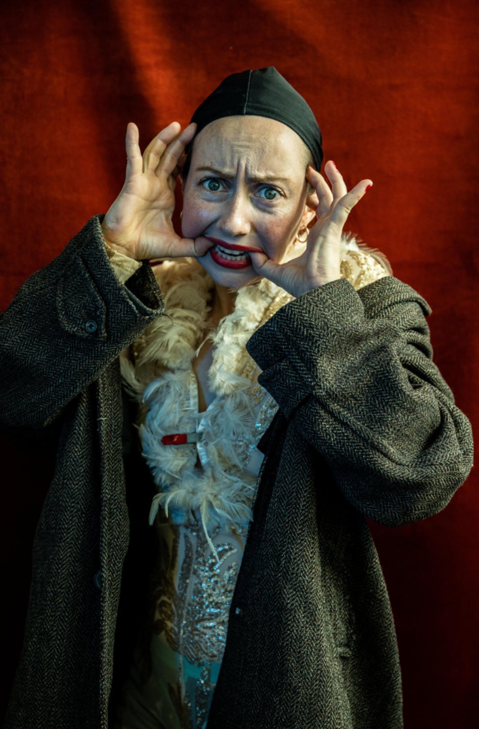 A performer with light skin poses with their thumbs in either side of their mouth in front of a red backdrop.