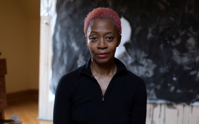 A woman with short pink hair sits barefoot on a cushion on a wooden floor. She is seated in front of a large black-and-white abstract artwork, with painting supplies scattered behind her.