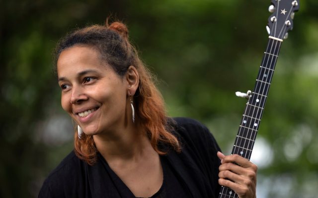 A woman with warm brown skin and copper-tinted hair smiles while holding a black banjo decorated with a star on its headstock.