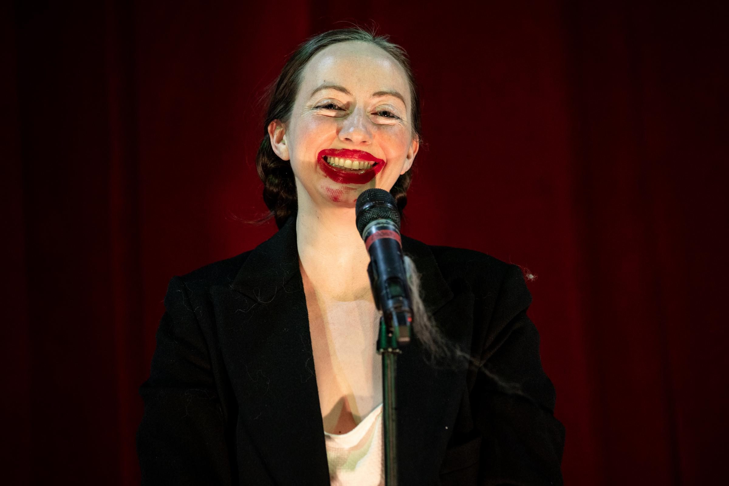 A performer with light skin smiles in front of a microphone, lipstick smeared all over their lips and chin.