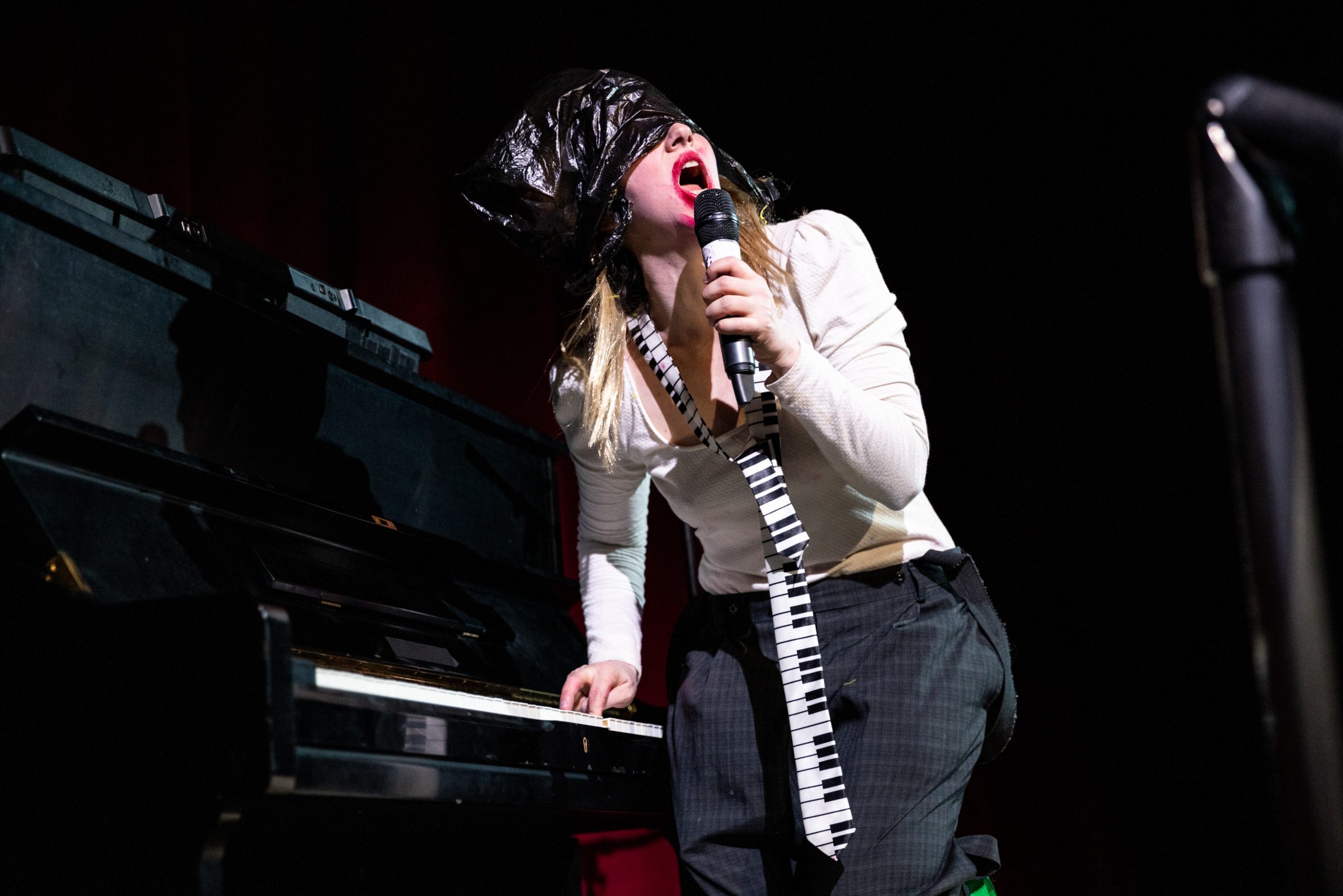A performer with light skin wearing a piano tie and black plastic bag over their head plays an upright piano.