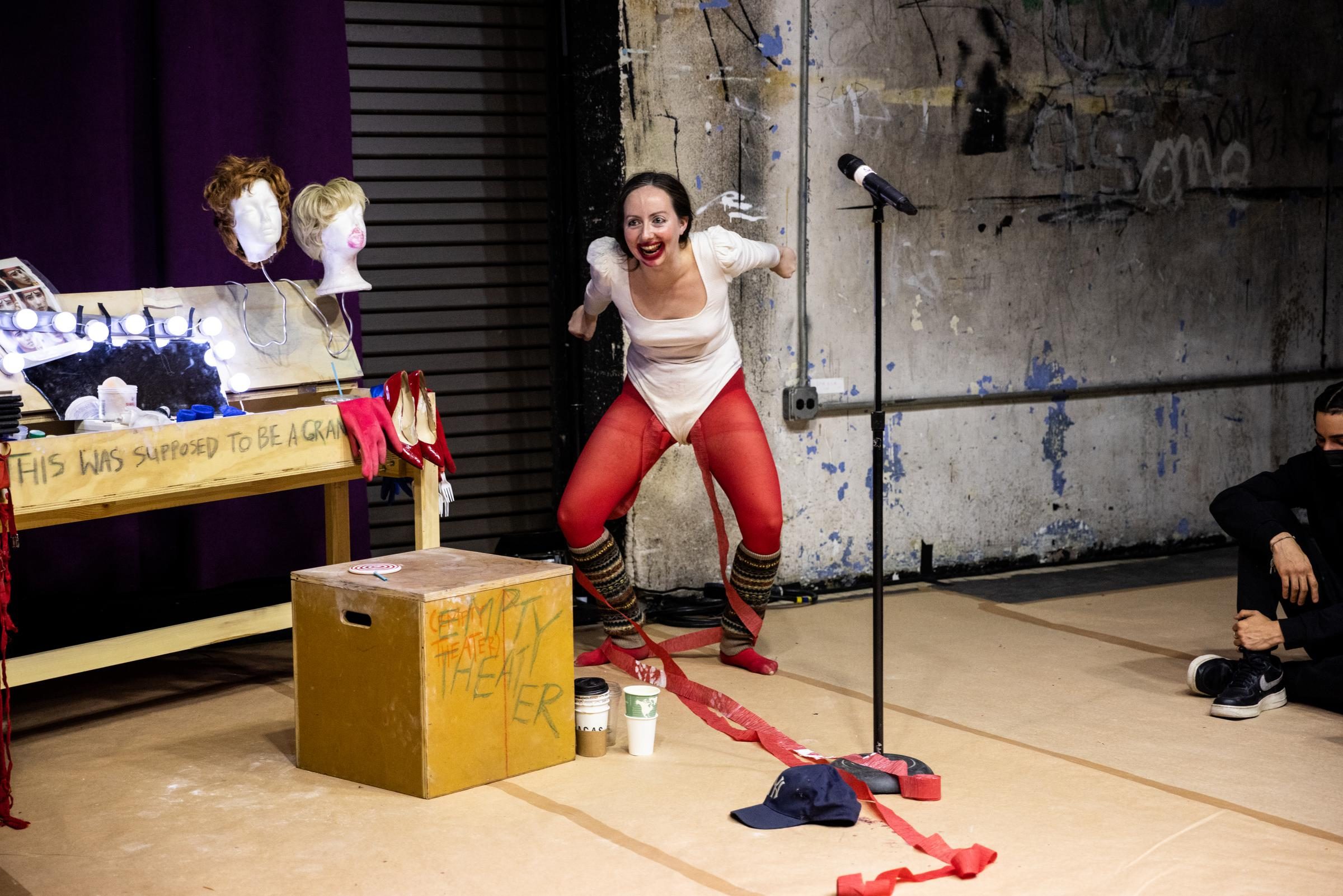 A performer with light skin with red streamers running from inside their white leotard prepares to jump onto a yellow box.