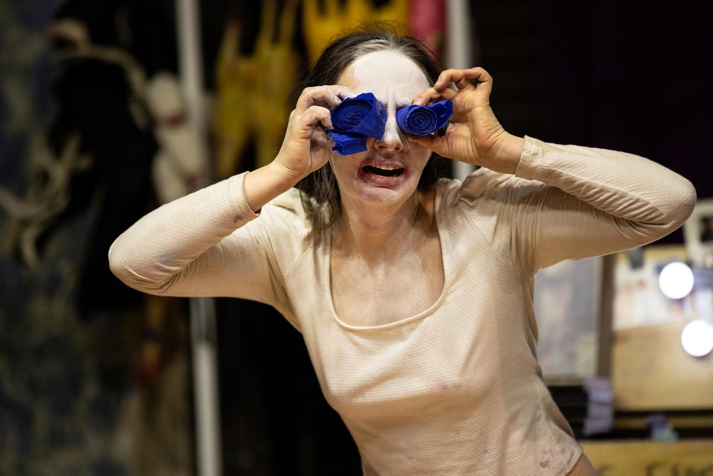 A performer with light skin with white paint on their face holds two rolls of blue streamers up to their eyes.